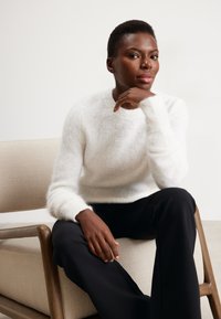 Pullover bianco e soffice in maglia con scollo rotondo e maniche lunghe, abbinato a pantaloni neri. Modella seduta su una sedia di tessuto chiaro.