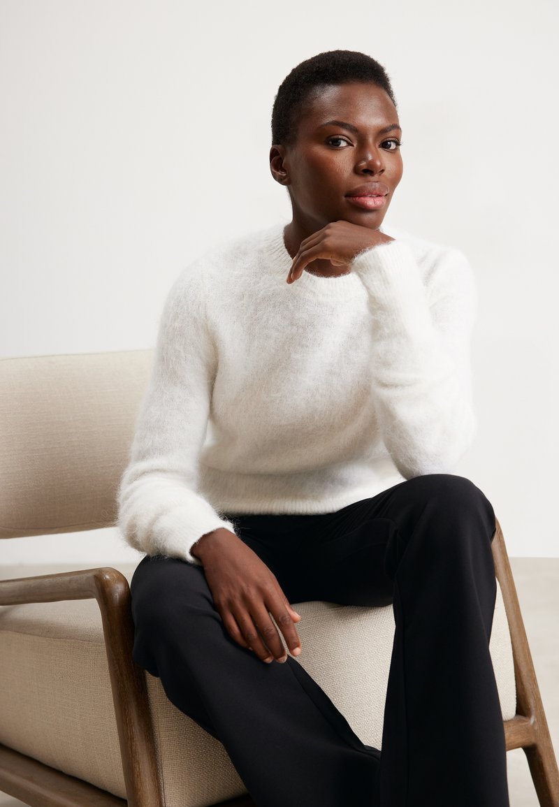 Pullover bianco e soffice in maglia con scollo rotondo e maniche lunghe, abbinato a pantaloni neri. Modella seduta su una sedia di tessuto chiaro.