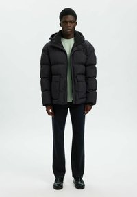 Mężczyzna stoi w czarnej kurtce puchowej, jasnoszarym swetrze, czarnych dżinsach i czarnych butach na tle jednolitego, jasnego tła.