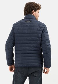 Marineblauwe gewatteerde jas met horizontaal gestikte patroon, hoge kraag en een klein logopatch op de rug. Draagt over donkere jeans.