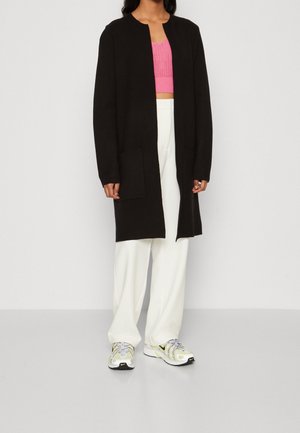 Persona con un largo cárdigan negro abierto, top de punto corto rosa, pantalones blancos de pierna ancha y zapatillas blancas y amarillas, de pie sobre un fondo liso.