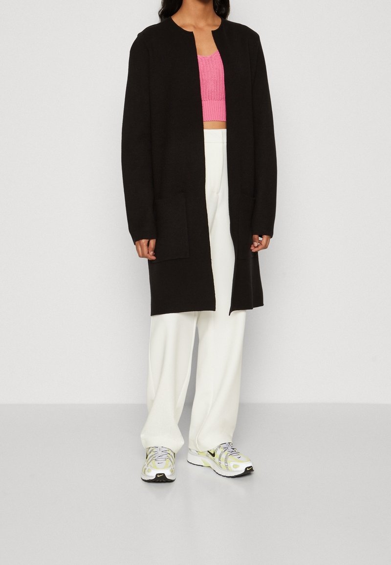 Personne portant un long cardigan noir ouvert, un top court en maille rose, un pantalon blanc à jambes larges et des baskets blanches et jaunes, debout sur un fond uni.