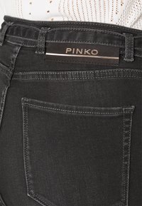 Svarta denimjeans med en slät textur, med en synlig bakficka och en märkesetikett med ordet "PINKO" fäst vid midjan.