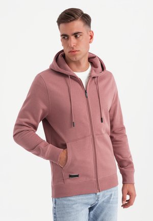 Młody mężczyzna w mauve kolorze bluzy z kapturem na zamek błyskawiczny z przednimi kieszeniami i białej koszuli, patrzący w lewo na jednolitym tle.