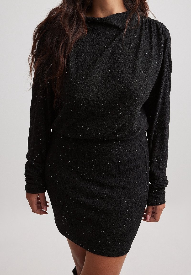 Femme portant une robe noire cintrée à manches longues avec des détails subtils scintillants et un col montant sur un fond uni.
