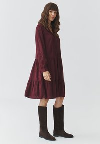 Robe bordeaux à volants avec des manches longues, présentant un motif de diamant subtil, associée à de hautes bottes en daim marron.