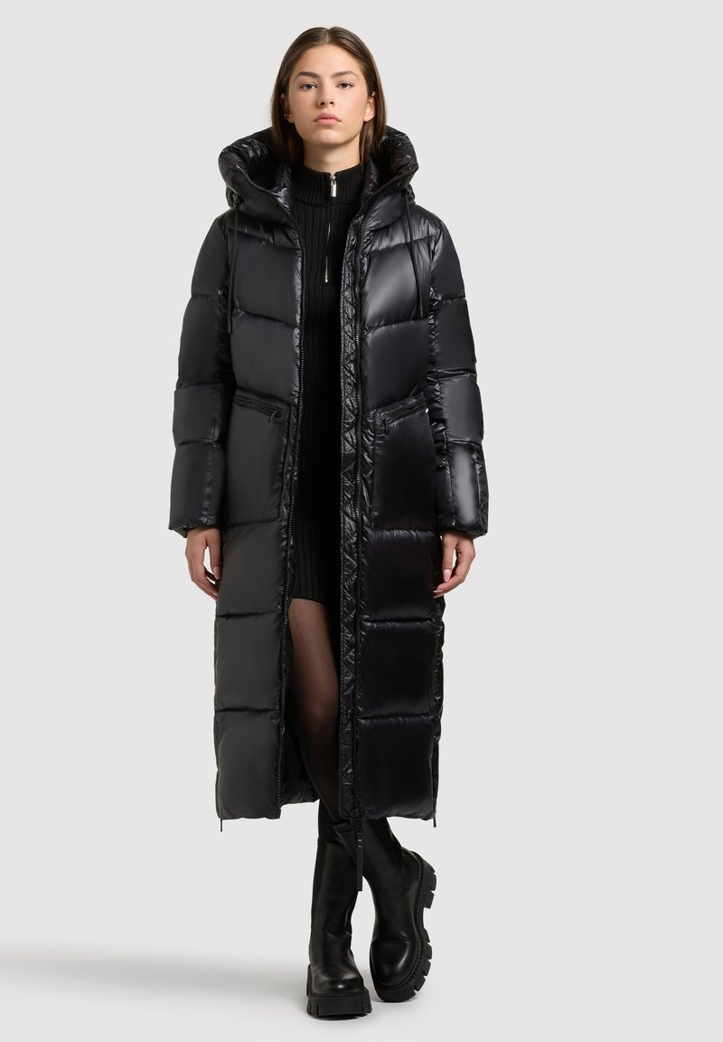 Zwarte pufferjas met een lange, oversized pasvorm. Kenmerken zijn een hoge kraag, twee zijzakken en een glanzende afwerking. Gecombineerd met chunky zwarte laarzen.