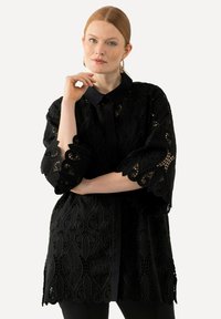 Túnica de encaje negro con estampados florales, con cuello tipo camisa y mangas tres cuartos. Presenta un corte recto y un dobladillo con forma de ondas.