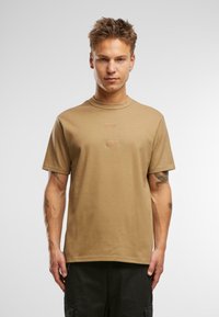 Brūns kokvilnas T-krekls ar īsām piedurknēm, apaļu kakla izgriezumu un diviem taisnstūrveida plāksteriem priekšpusē. Audums izskatās gluds, ar minimālu tekstūru.