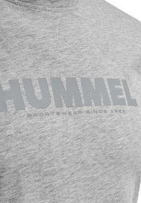 Pelēka kokvilnas T-kreklu ar apaļu kakla izgriezumu, uz kura ir liela pelēka teksta uzraksts "HUMMEL" un mazāks uzraksts "SPORTSWEAR SINCE 1923" zem tā.