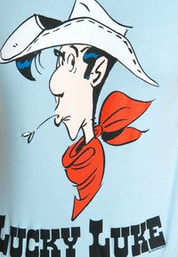 Light blue t-shirt featuring a cartoon character with black hair, white cowboy hat, and red bandana, text reads "LUCKY LUKE" in bold black letters.