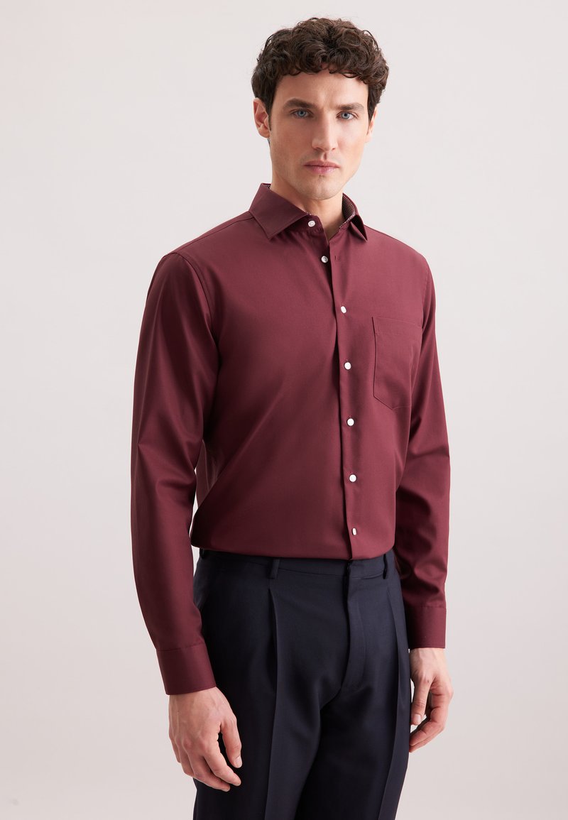 Uomo con camicia a maniche lunghe bordeaux con taschino e pantaloni scuri blu navy, in piedi su uno sfondo chiaro e semplice.