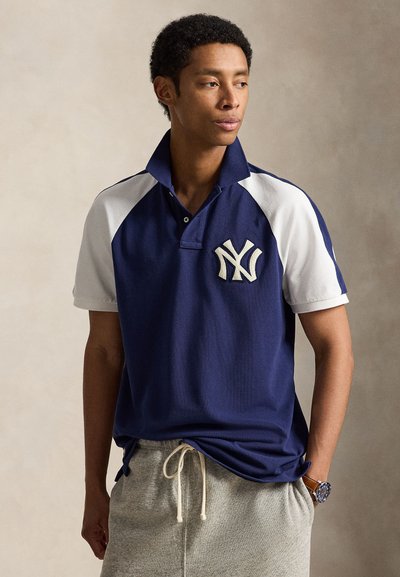 Mees kannab tumesinist ja valget lühikeste varrukatega polo särki NY logoga, halli paelaga pükse ja kella, vaadates küljelt ühetaustalise tausta vastu.