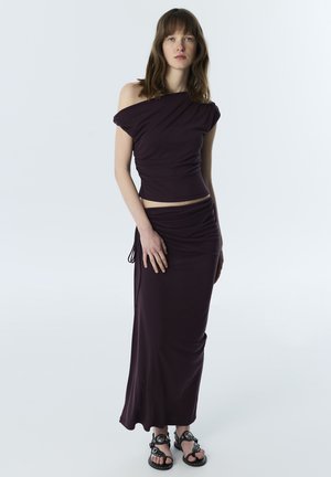 Junge Frau steht und trägt ein dunkelviolettes One-Shoulder-Top und einen passenden langen Rock mit schwarzen, verzierten Sandalen vor hellem Hintergrund.
