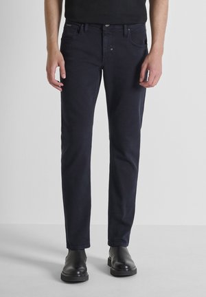 Homme portant un jean noir ajusté et des bottes en cuir noires, se tenant contre un fond gris clair uni.