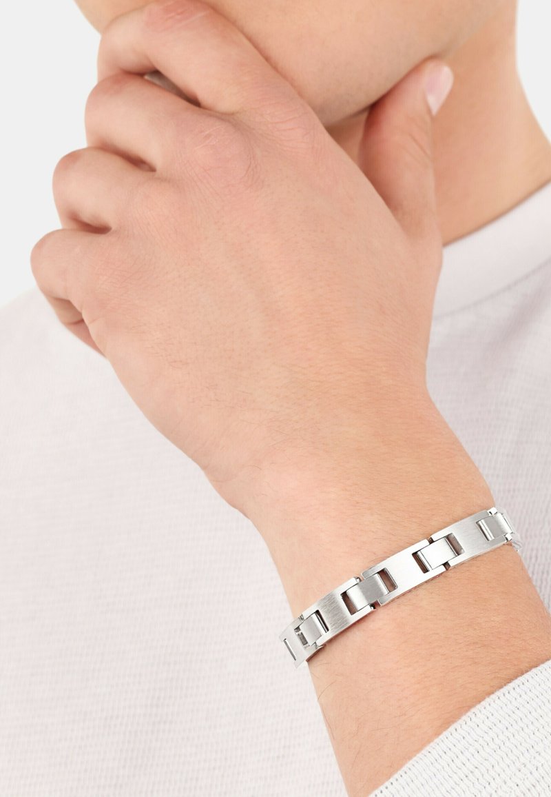 Pulsera de metal plateado con eslabones rectangulares entrelazados y acabado cepillado. Lleva puesta en una muñeca, complementando una camisa de color claro.