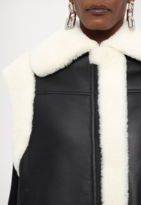 Gilet en cuir noir avec un col en fausse fourrure blanche. Présente un design épuré, des coutures visibles et une fermeture éclair à l'avant. Boucles d'oreilles en chaîne notables.