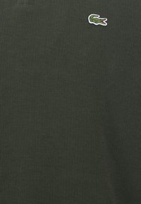Tamsiai žalia šonkaine megzta polo marškinėliai su mažu siuvinėtu krokodilo logotipu baltos ir žalios spalvų ant kairiojo krūtinės.