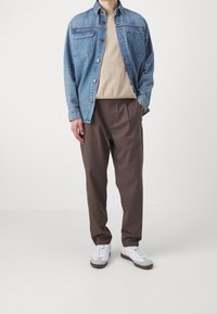 Denimjack, lichtblauw, met borstzakken; beige trui eronder; bruine plooibroek; witte sneakers met grijze accenten.