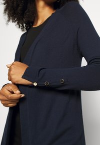 Námořnická modrá pletená kardigan s šálovým límcem, slim fit a zlatými knoflíkovými akcenty na rukávech. Měkká textura a neformální design.