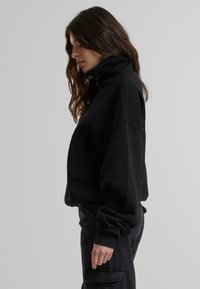 Svart fleece genser med høy krage, senket skuldre, og mansjetter ved ermene. Glatt tekstur; oversized design.