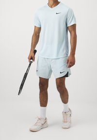 Hellblaues Nike V-Ausschnitt-T-Shirt, passende Shorts mit schwarzen Akzenten, hält einen schwarzen Tennisschläger und trägt weiße Sneaker mit pinken Details.