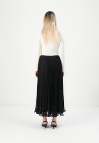 Mulher com cabelo ombre, vestindo uma blusa branca de manga longa, uma saia maxi plissada preta e sandálias de salto pretas, posicionada contra um fundo branco.