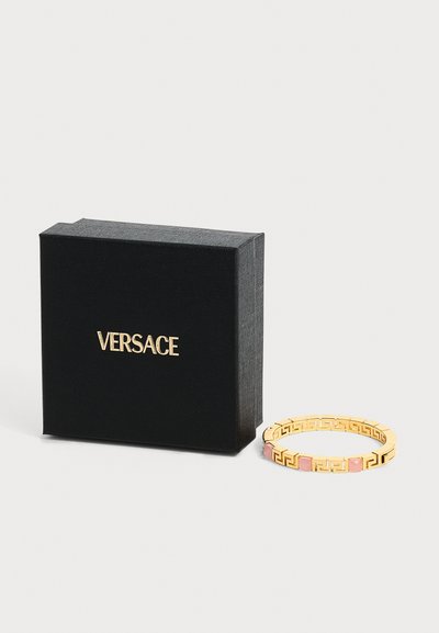 Μαύρο κουτί Versace με χρυσό λογότυπο δίπλα σε χρυσό βραχιόλι με ροζ τετράγωνες πέτρες και γεωμετρικό σχέδιο με κοψίματα.