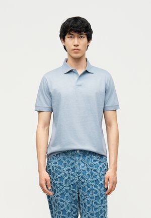 Bărbat purtând o tricou polo cu mâneci scurte, de culoare albastru deschis, și blugi albaștri cu un model floral în relief, stând în fața unui fundal alb simplu.