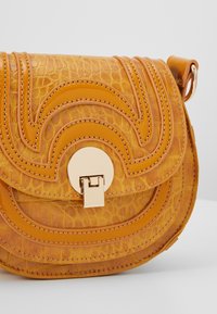Piccola borsa a tracolla color beige con texture a coccodrillo, cuciture curve sovrapposte e un fermo rotondo dorato su uno sfondo chiaro.
