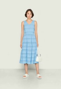 Vestido azul claro, sem mangas, com corpete ajustado e saia rodada. Apresenta riscas horizontais e detalhes em ilhós. Usado com sandálias brancas e uma bolsa.