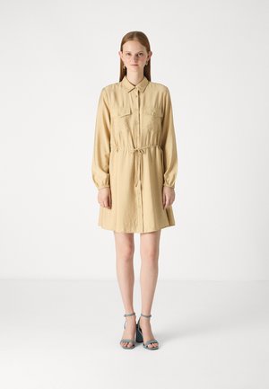 Robe chemise beige à boutons avec deux poches poitrine, une taille élastique et des manches longues. Associée à des talons à bride de cheville bleus.