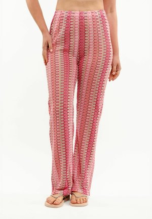 Kobieta w różowych, wzorzystych spodniach z wysokim stanem o prostych nogawkach i beżowych sandałach, stojąca na jednolitym białym tle.