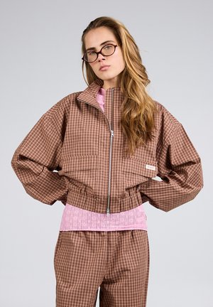 Frau mit langen Haaren und Brille trägt eine braun karierte Jacke und Hose über einem rosa Spitzenoberteil, steht mit Händen in die Hüften gestützt vor einem schlichten Hintergrund.