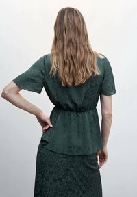 Ciemnozielona bluzka z krótkimi rękawami, luźnym krojem i marszczonym w talii pasem. Tkanina ma subtelny kwiatowy wzór i gładką fakturę.