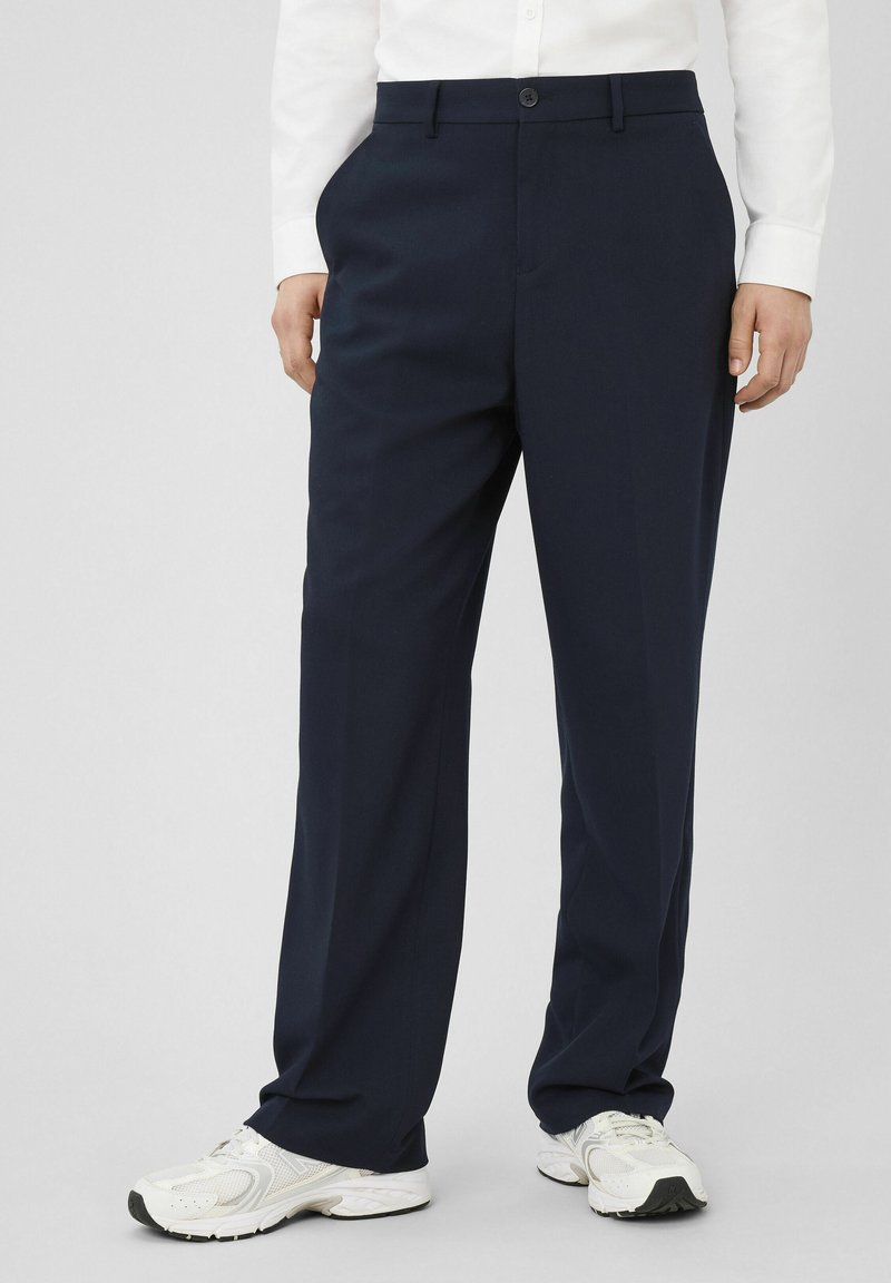 Personne portant un pantalon bleu marine sur mesure, une chemise blanche à manches longues et des baskets blanches, debout devant un fond uni.