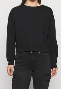 Sudadera negra de manga larga, con cuello redondo, dobladillo y puños acanalados, hecha de material de algodón suave. Lleva pantalones cargo negros.