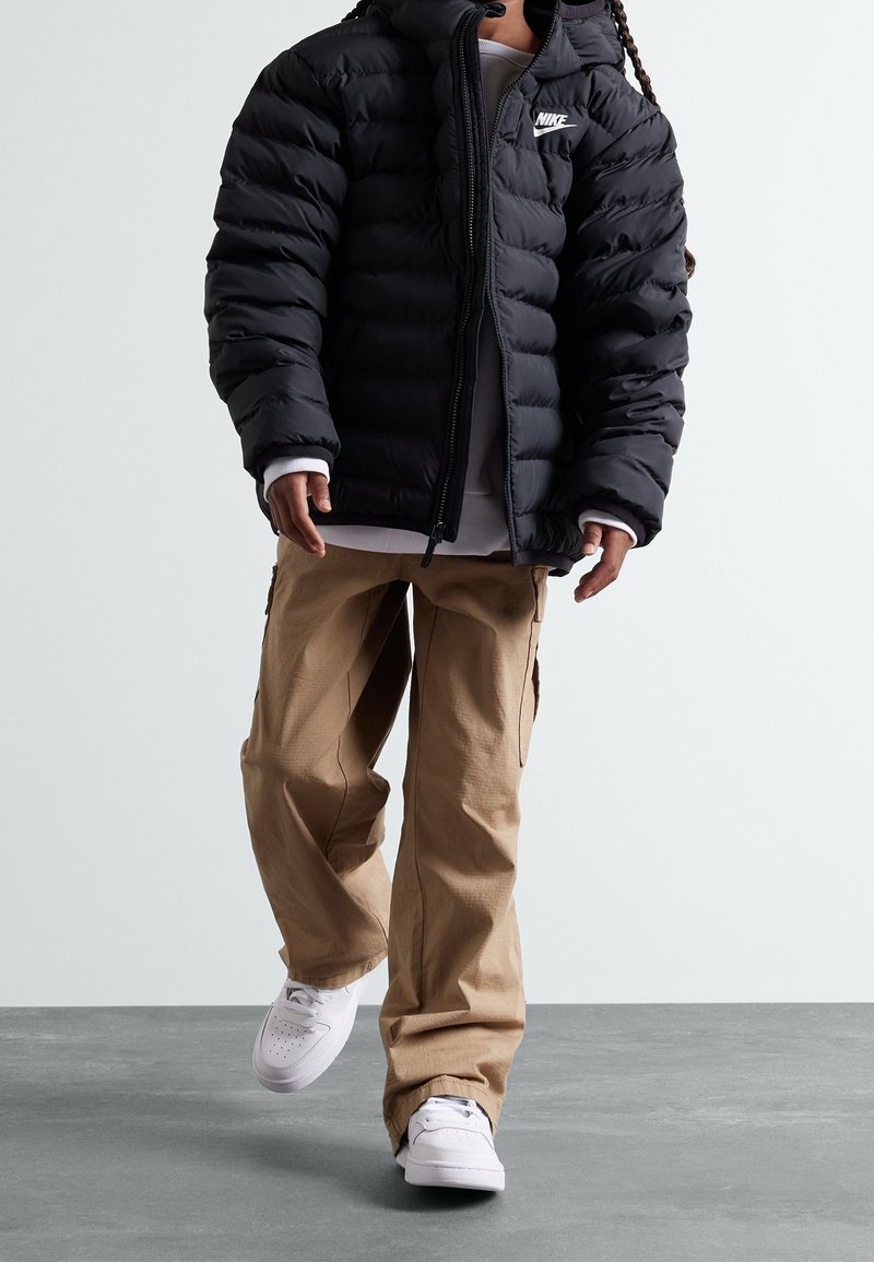 Chaqueta de plumas negra con cremallera y logo de Nike, pantalones cargo beige y zapatillas blancas, que presentan un ajuste relajado y una tela texturizada.