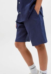 Kā navy zila drupinātā auduma šorti ar sānu kabatām, līdz ceļgaliem, apvienoti ar baltām zeķēm ar melnu tekstu. Tiek attēlots, ka tekstūra ir mīksta un viegla.