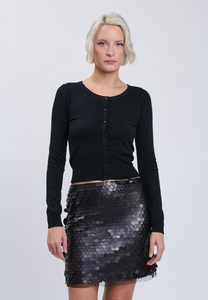 Vrouw met kort blond haar draagt een zwarte geknoopte cardigan en een glanzende zwarte pailletten minirok, staand tegen een effen lichte achtergrond.
