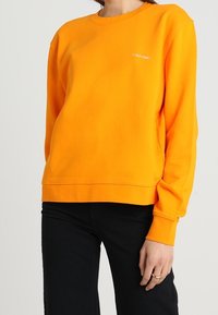 Pomarańczowa bawełniana bluza z okrągłym dekoltem, z prążkowanym kołnierzem i mankietami oraz małym białym logo na lewej piersi. Nosi się ją z czarnymi spodniami.