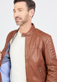 Veste en cuir marron avec fermeture éclair, dotée de détails côtelés aux épaules et d'une doublure à carreaux bleus. Le modèle porte une chemise claire en dessous.