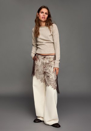Jasnobeżowy dzianinowy sweter z długimi rękawami, zestawiony z szerokimi kremowymi spodniami z ciemną koronkową nakładką i kwiatowym wzorem u dołu.