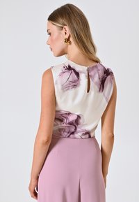 Blusa senza maniche bianca con stampa floreale viola, caratterizzata da un'apertura a chiave sul retro e una texture morbida. Abbinata a una gonna rosa.