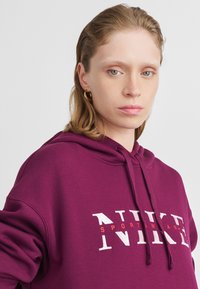 Maroon kapuco s puloverjem z belim in rdečim besedilom "NIKE SPORTSWEAR". Narejen iz mehkega materiala z nastavljivimi vrvicami in sprednjo torbico.