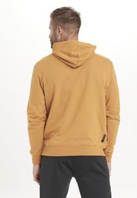 Sennepsgul hoodie med snorhætte, ribbede ærmer og kant. Lavet af blødt materiale, har en lille sort label på den nedre side.