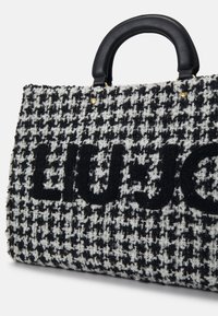Czarna i biała torebka w kratę psią z teksturowanej tkaniny o splocie, z czarnymi skórzanymi rączkami i wyraźnym haftowanym logo. Złote metalowe detale.