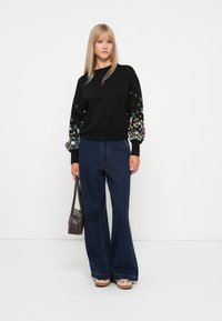 Felpa nera con maniche ricamate a fiori, abbinata a jeans a gamba larga blu. Il modello porta una borsa a tracolla marrone scuro e indossa scarpe beige.