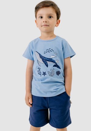 Young boy wearing a light blue shirt with a whale and fish prints, paired with dark blue shorts, standing with one hand in pocket.