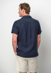 Camicia navy a maniche corte in tessuto di lino, con taglio dritto, colletto a punta e una texture sottile. Indossata con pantaloni corti chiari.